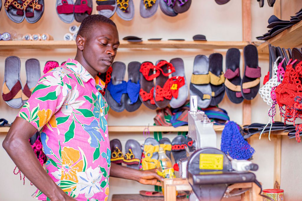 A craftsman wearing a floral shirt working in a shoe shop surrounded by handmade sandals.
